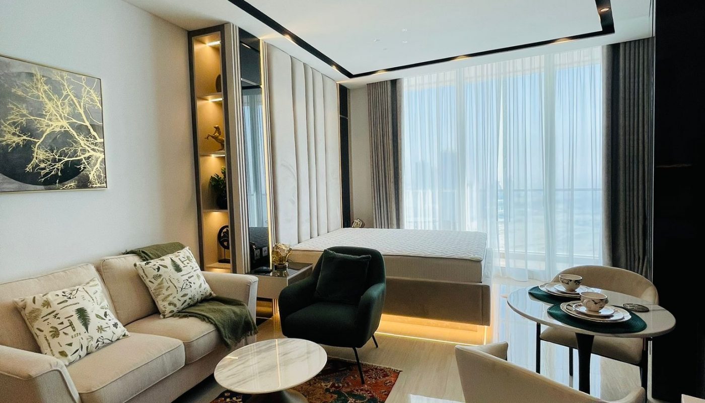 Modern studio apartment with a bed, sofa, armchair, round coffee table, small dining table, and large windows with sheer curtains allowing natural light.