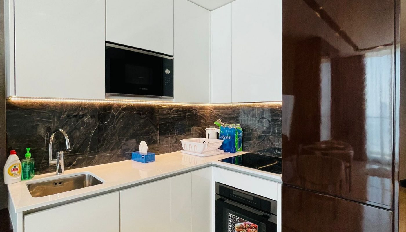 Modern kitchen with white cabinets, a dark backsplash, built-in appliances, a brown refrigerator, and a rug on the floor. Various cleaning products are near the sink.