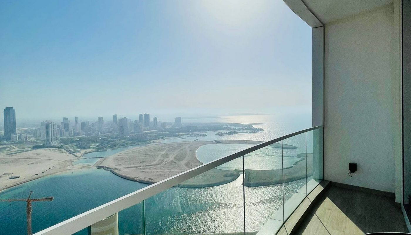 High-rise balcony with glass railing overlooks a city skyline, sandy coastline, and calm water under a bright sun. A glass-top table and chair are visible in the corner.