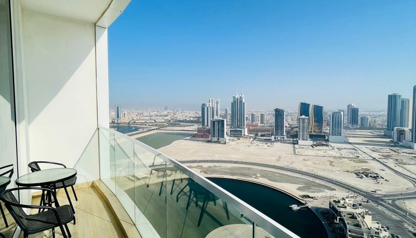 High-rise balcony with glass railing, small table and two chairs, overlooking a modern cityscape with tall buildings and a waterway under a clear blue sky.
