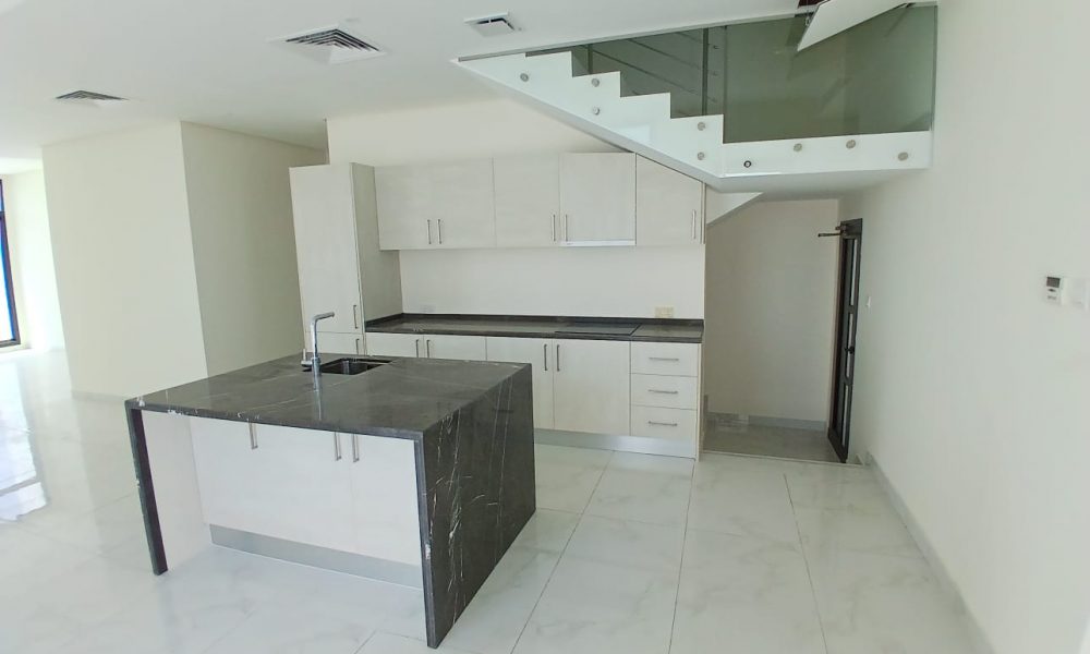 Luxurious modern kitchen with a central marble island, white cabinets, and a stainless steel sink in a Modern Villa. A staircase with glass railing is above the kitchen area. The room boasts white walls and a tiled floor, offering an exquisite feature reminiscent of Diyar Al Muharraq's design elegance.