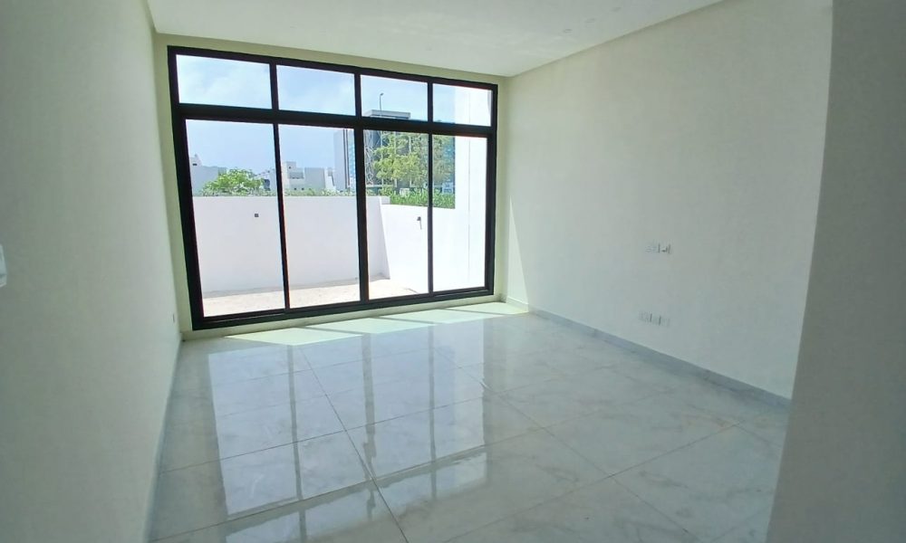 A luxurious, vacant, well-lit room with large floor-to-ceiling windows overlooking a small terrace in Diyar Al Muharraq. The space boasts white walls and a light-colored tiled floor, perfect for adding a touch of elegance to any modern villa.