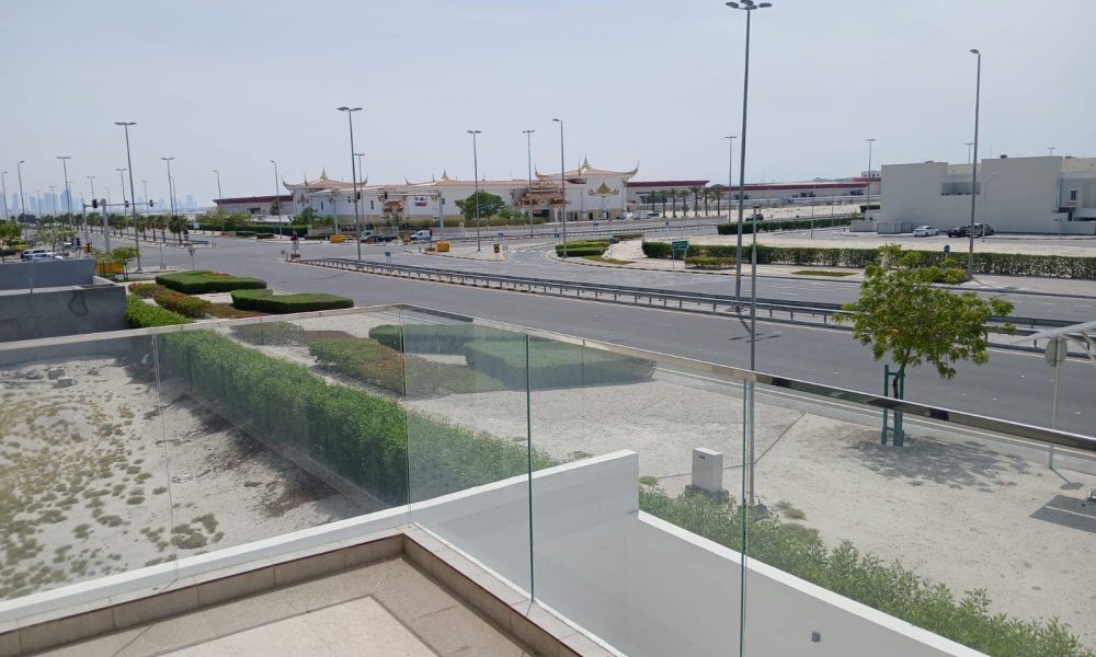 View from a building's balcony showing a multi-lane road with light traffic, green shrubs, and trees. The city buildings, including a luxurious modern villa in the distance, display distinct architectural roofs against the backdrop of a clear sky.