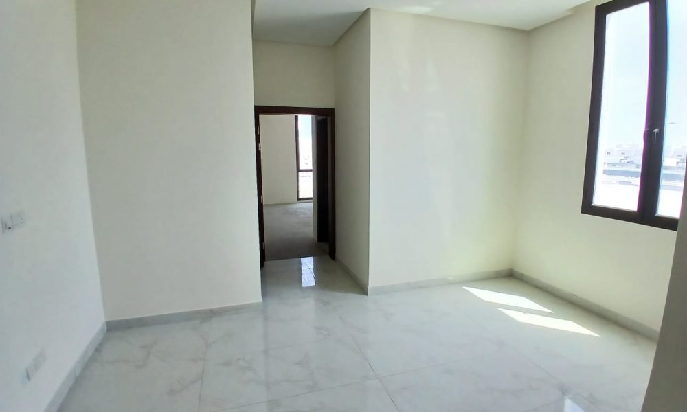 A bright, empty room with white walls and a tiled floor in a luxurious modern villa at Diyar Al Muharraq, featuring a single large window on the right and a doorway leading to another similarly empty room.