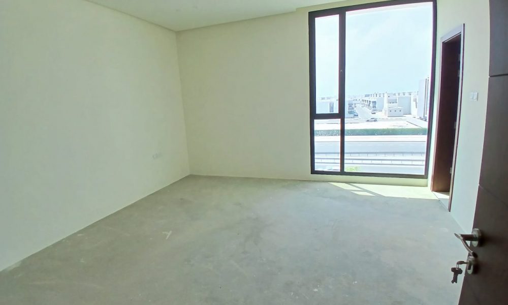 A vacant, unfurnished room in a luxurious modern villa with off-white walls, a large window offering views of the buildings in Diyar Al Muharraq, and no flooring or ceiling fixtures.