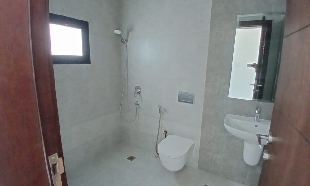 A minimalist bathroom in a modern villa at Diyar Al Muharraq, with a wall-mounted toilet, sink, faucet, and showerhead. It features luxurious grey tiled walls and a small window above the shower.