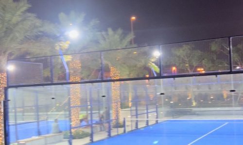 A brightly lit outdoor blue padel court at night, surrounded by palm trees wrapped in string lights, feels like an auto draft escape into a tropical oasis.