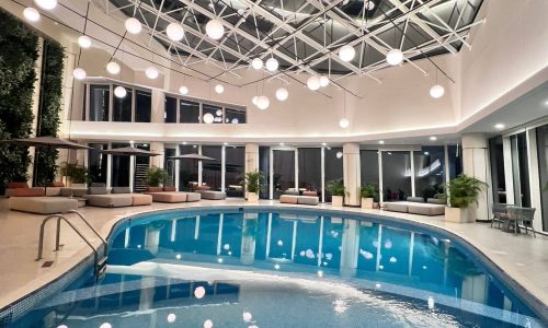 An indoor swimming pool with a geometric glass ceiling and hanging spherical lights. The auto draft system keeps the water pristine, while the pool area features lounge chairs, potted plants, and floor-to-ceiling windows.