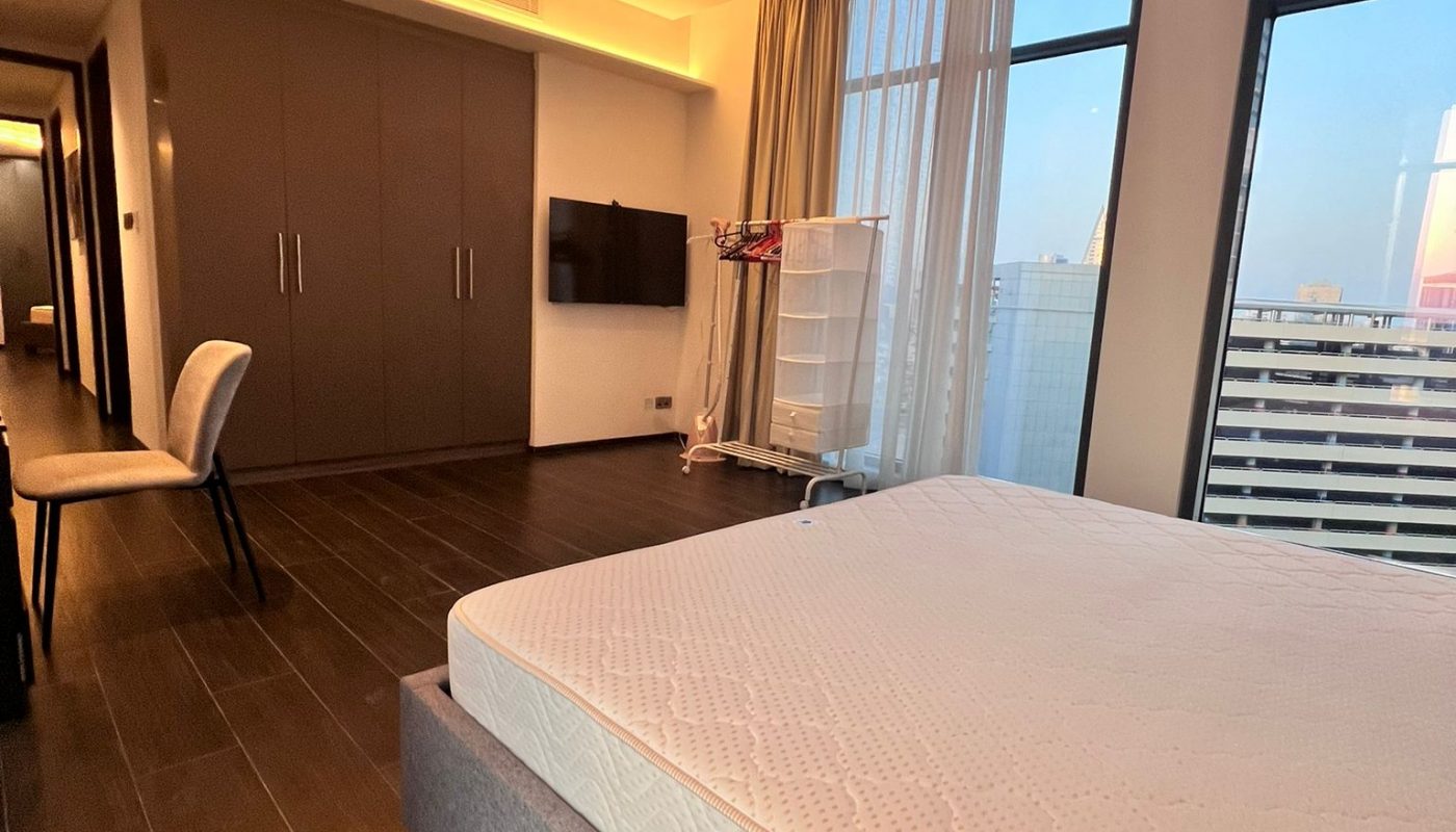 Modern bedroom with a large bed, wall-mounted TV, wardrobe, chair at a desk, and large windows letting in natural light.
