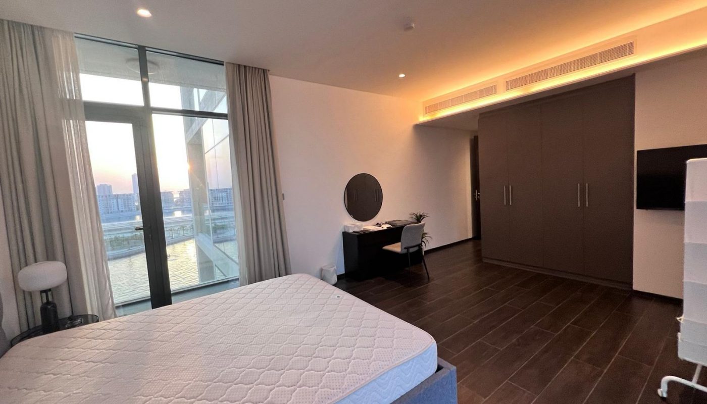 Modern bedroom with large window, unmade bed, built-in wardrobe, desk with chair and round mirror, and a city view at sunset.