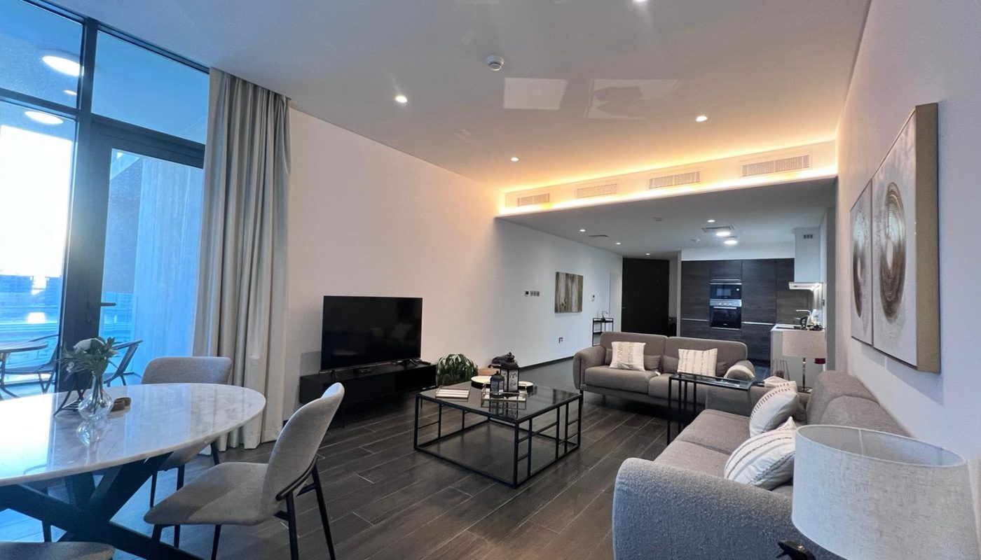 Modern living room with gray sofas, a TV, a round dining table with chairs, and an open kitchen area. Large window lets in natural light.