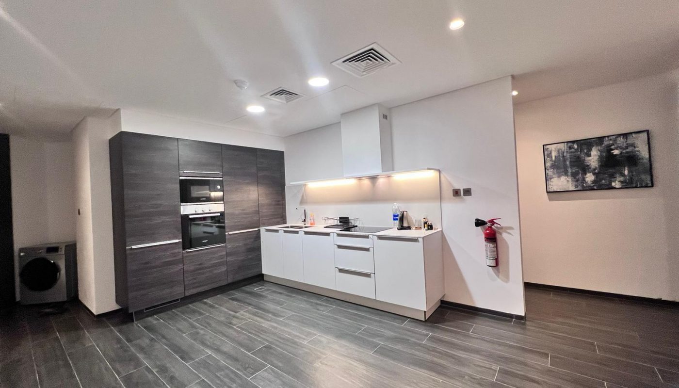 Modern kitchen with dark and white cabinets, built-in oven, stovetop, under-cabinet lighting, fire extinguisher on the wall, and a washing machine in the corner.