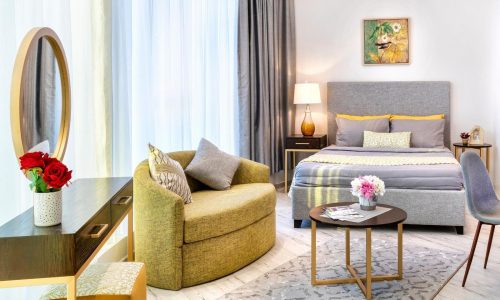 A modern studio for rent in the Seef area features a gray bed with yellow accents, a round chair, and a wooden desk with a mirror. The space includes a small round coffee table and is decorated with flowers, a painting, and floor-to-ceiling curtains.