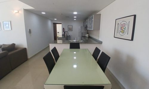 A modern dining area with a glass-top table and six black chairs is shown in this Amwaj flat for rent. In the background, there is an open kitchen with granite countertops and white cabinets. Art is hung on the wall to the right.