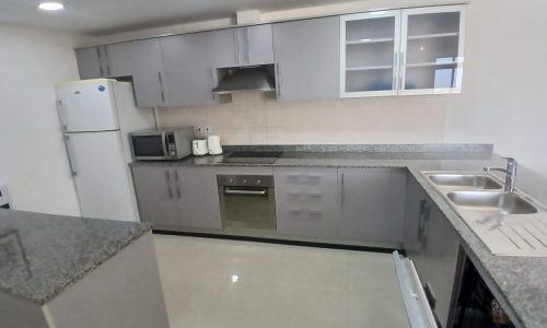 A modern kitchen in an Amwaj flat for rent, equipped with stainless steel appliances including an oven, microwave, and refrigerator. The room features grey cabinetry, a double sink, and a granite countertop, with a partially open dishwasher.