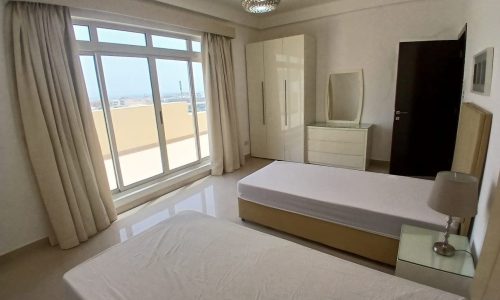 A clean, minimalistic bedroom in an Amwaj flat for rent features two single beds, a wardrobe, a dresser with a mirror, and a lamp on a bedside table. A large window with curtains opens to a view outside.