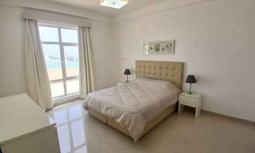 A minimalist bedroom in an Amwaj flat for rent features a large bed, two bedside tables with lamps, a dresser, and a large window with white curtains.