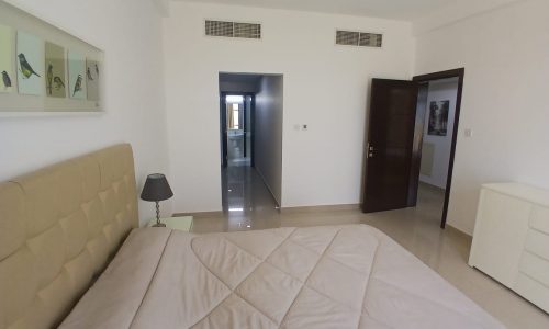 A bedroom in the Amwaj flat for rent features a beige upholstered bed, a nightstand with a lamp, and a dresser. A hallway leads to another room. The white walls are adorned with a bird-themed picture frame above the bed.