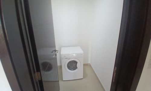 A small, white laundry room in an Amwaj flat for rent features a single front-loading washing machine against the back wall. The room has beige tiled flooring and white walls, with a black door frame partially visible.