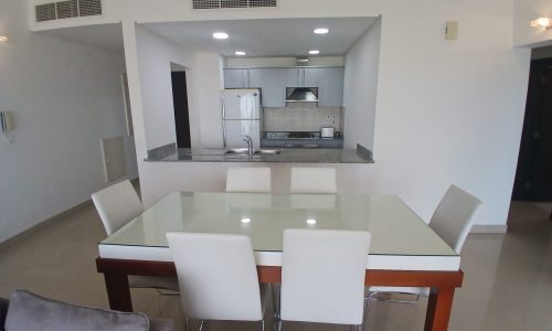 A modern kitchen and dining area in a spacious apartment on Amwaj Islands features a glass-top dining table with six white chairs. The kitchen boasts white cabinets, a stainless steel refrigerator, and a built-in stove and oven, all complemented by an awe-inspiring sea view.