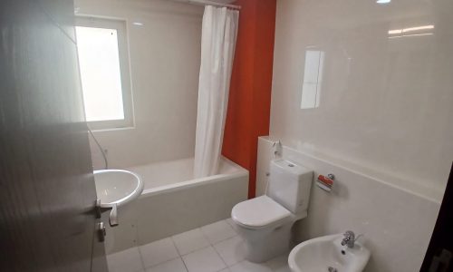 A bathroom in a spacious apartment with a bathtub, shower curtain, sink, toilet, and bidet. The walls are white with one side featuring orange tiles, and there is a window above the bathtub offering a partial sea view of the Amwaj Islands.