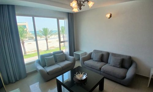 A living room in a stunning rental property in Muharraq features two blue-gray sofas, a black coffee table with a bowl, and a large window showcasing a beach view with palm trees. This flat for rent offers an inviting and serene atmosphere perfect for relaxation.