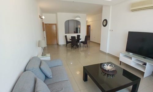 A modern flat for rent in Muharraq features a living room with a gray sofa, black coffee table, flat-screen TV, dining table with four chairs, clock on the wall, and air conditioner. A light fixture and doorway are visible in the background.