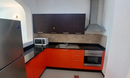 A compact kitchen in a Muharraq flat for rent features red lower cabinets, dark upper cabinets, stainless steel appliances including a refrigerator, microwave, and oven, a sink, and a range hood. The tiled backsplash adds a perfect finishing touch.