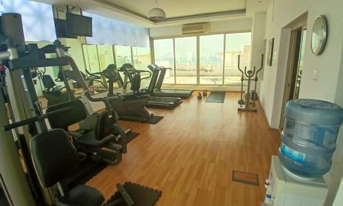 A gym room with exercise equipment including treadmills, ellipticals, and a water cooler is available in this Muharraq apartment. The room features wooden flooring and large windows allowing natural light—a perfect amenity when you rent in Muharraq.