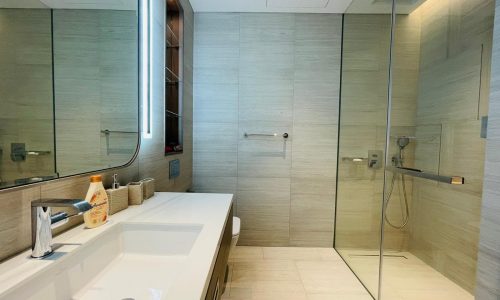 A modern apartment bathroom in Bahrain Bay features a large mirror, sink, soap dispenser, and glass-walled shower. The tiled walls and floor add a sleek touch, complemented by a towel rack on the wall. Perfect for rent!