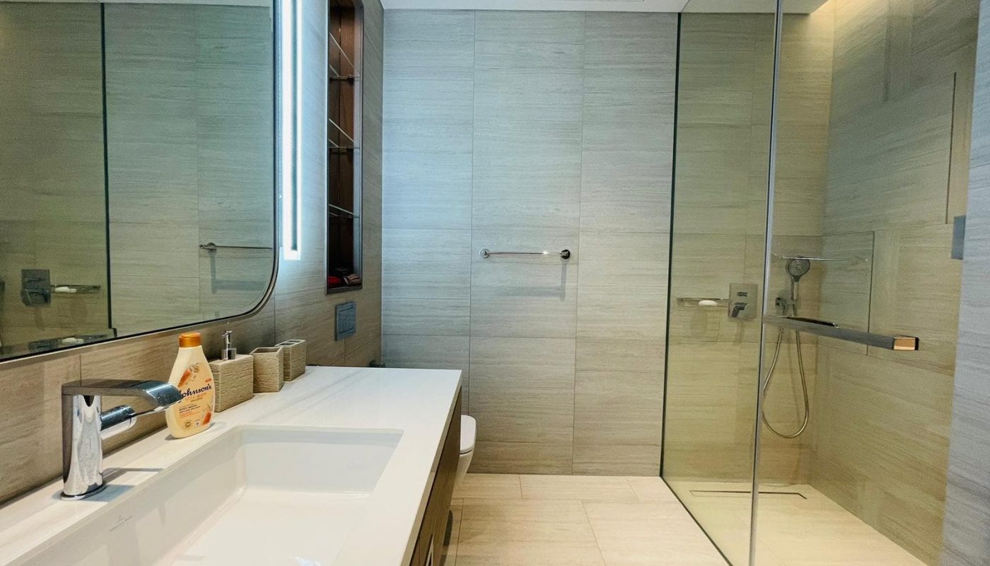 A modern apartment bathroom in Bahrain Bay features a large mirror, sink, soap dispenser, and glass-walled shower. The tiled walls and floor add a sleek touch, complemented by a towel rack on the wall. Perfect for rent!