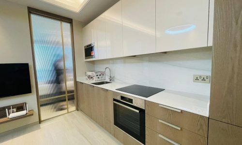 Modern kitchen with a white and wood-toned design. Cabinetry includes integrated appliances like an oven, microwave, and cooktop. A sliding door and flat-screen TV are visible in the corner of this stylish Bahrain Bay apartment for rent.