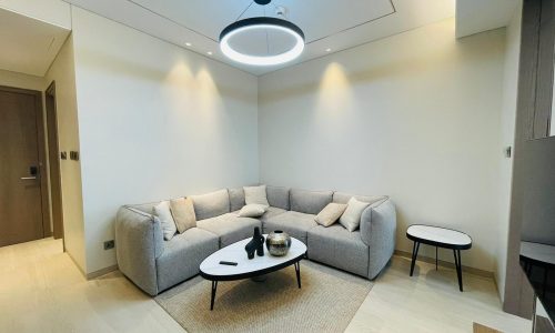 A modern living room in an apartment for rent in Bahrain Bay, featuring a light gray sectional sofa, white oval coffee table, round side table, and a central ring light. The walls are white, and the floor is light wood.