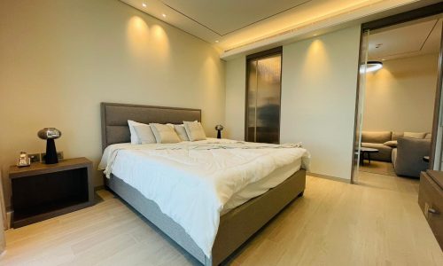 Modern apartment bedroom with a large bed, white bedding, and two small bedside tables with lamps. There's a partially visible adjoining sitting room with a couch in the background, perfect for those looking to rent in Bahrain Bay.