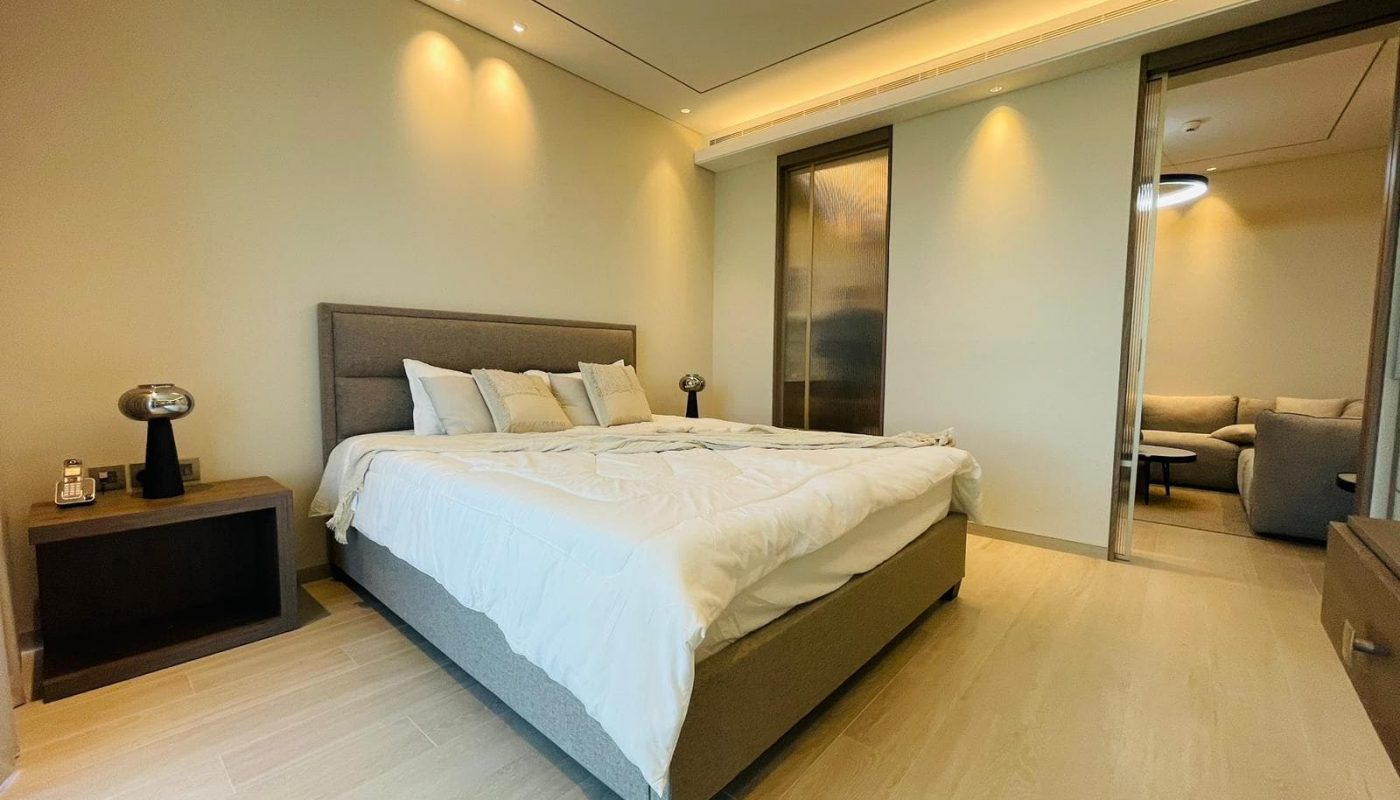 Modern apartment bedroom with a large bed, white bedding, and two small bedside tables with lamps. There's a partially visible adjoining sitting room with a couch in the background, perfect for those looking to rent in Bahrain Bay.