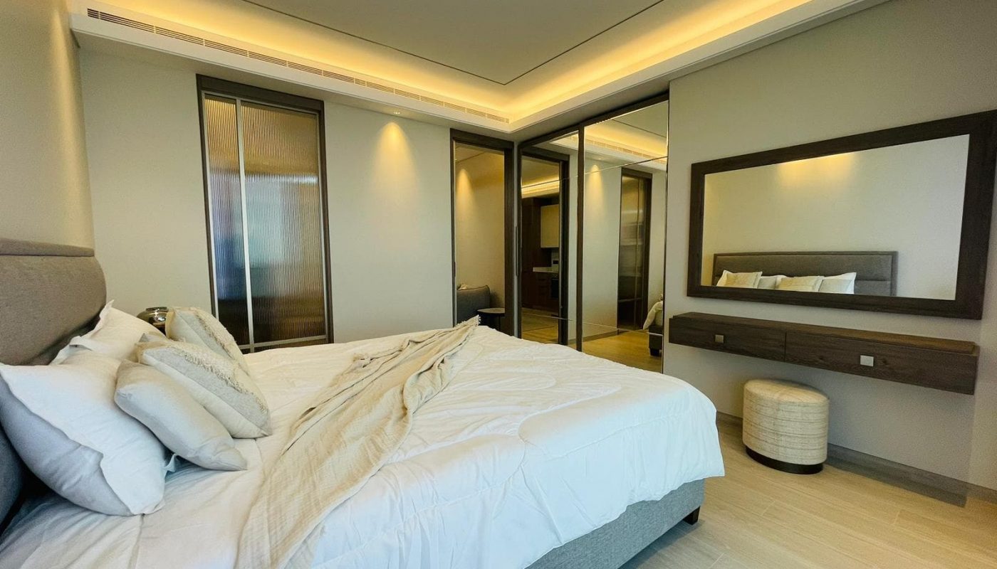 Modern apartment bedroom in Bahrain Bay with a neatly made bed featuring white linens, mirrored sliding closet doors, a wooden vanity with a large mirror, and ambient ceiling lighting. Perfect for rent!