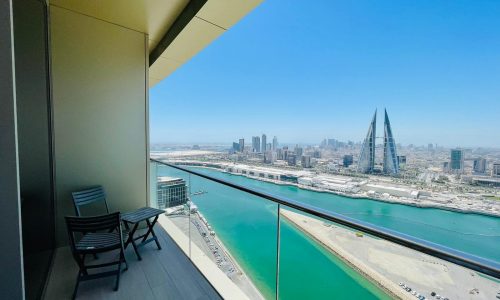 A rented apartment features a balcony with chairs and a table, overlooking the stunning cityscape of Bahrain Bay. The view includes tall buildings, a body of water, and the distinctive twin-towered skyscraper.