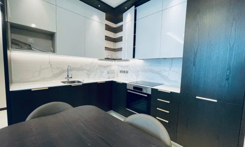 Modern kitchen in a Seef apartment with white upper cabinets, dark lower cabinets, marble backsplash, built-in oven, sink, and induction cooktop. Dark dining table and chairs in the foreground. Available for rent now.