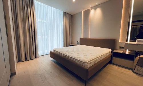 A modern bedroom in an apartment for rent with a double bed, beige curtains, and light wooden flooring. The Seef room includes a bedside table, a wall-mounted light, and a large window with sheer curtains.