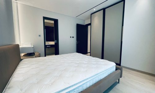 A bedroom in a spacious Seef area apartment for rent, featuring a large bed, a built-in wardrobe with sliding doors, and a view of an adjacent bathroom. The room is well-lit with modern, minimalist decor.