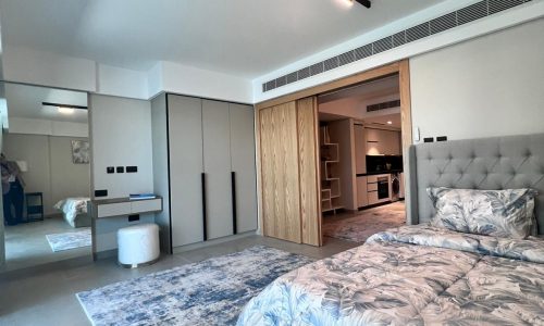 A modern bedroom with a grey upholstered bed, floral bedding, a large mirror, and sliding wooden closet doors. A blue rug is on the floor, and the room has recessed lighting.