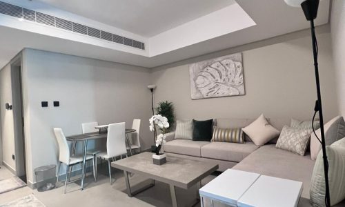 A modern, neutral-toned living room and dining area with a sectional sofa, coffee table, dining table with chairs, floor lamp, and wall art. Decor includes cushions and a plant, creating a cozy atmosphere.