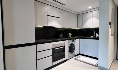 A modern kitchen with white cabinets, black countertops, built-in oven, washing machine, and small appliances on the counters, illuminated by under-cabinet lighting.