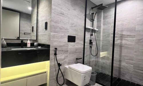 Modern bathroom with grey tiles, a wall-mounted toilet, black fixtures, and a walk-in glass shower. An illuminated vanity with a large mirror adds elegance, complemented by two grey floor mats. This auto-draft design seamlessly blends function and style for a contemporary feel.