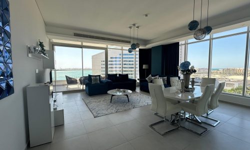 A modern living and dining area in this Amwaj rental property features a blue sofa, a white dining table, and large windows offering city and water views. Decorative blue and gray accents are present throughout the flat for rent.