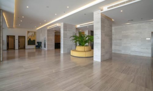 Spacious, modern lobby with beige marble floors, white walls, and several large columns. A round yellow seating area with a large plant in the center is present in the middle of the space, perfect for welcoming guests to your Amwaj flat for rent.