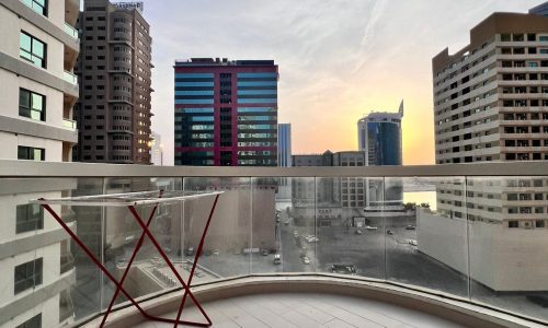 A balcony in a Juffair apartment overlooking a cityscape with a mix of buildings at sunset. There is a red metal chair positioned against the glass railing on the tiled floor, offering a serene spot to relax for those lucky enough to rent this view.