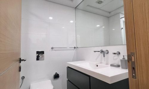 A modern bathroom in a Juffair apartment with a wall-mounted toilet, a double-sink vanity, large mirror, and white tiled walls. The floor has dark mats and a towel rail mounted on the wall, perfect for comfortable living when you rent in this vibrant area.