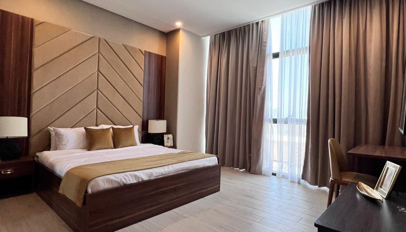 Modern hotel room with a large bed, bedside tables, a desk with a chair, and floor-to-ceiling windows covered by curtains. The room features wood accents and light-colored flooring.