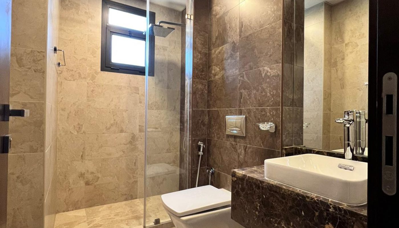 Modern bathroom with beige marble floors and walls, glass-enclosed shower, wall-mounted toilet, bidet spray, and a rectangular sink set in a brown marble countertop.
